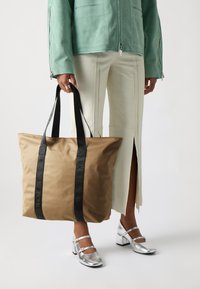 Barna bevásárlótáska fekete fülekkel, texturált anyagból készült, logókiemelésekkel. A modellen világos színű, slim-fit nadrág és ezüstcipők láthatók.