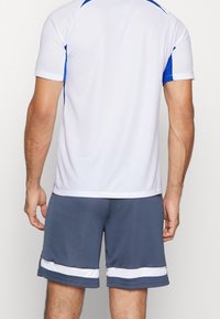 Camiseta atlética blanca con acentos azules, combinada con pantalones cortos grises que presentan rayas blancas. La tela parece suave y ligera.