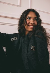 Schwarze Kapuzenjacke mit weicher Textur; mit goldenen gestickten Akzenten eines Sterns und dem Text "LUCKY STAR". Einfachheit im Design, funktionale Form.