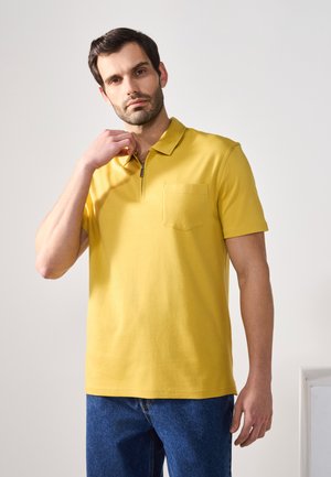 Gul polo-shirt med lynlås i halsen og forlommer, lavet af glat bomuldsstof. Iført med blå jeans, med korte ærmer og lige kant.