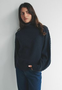 Tmavomodrý rolákový sveter z mäkkej úpletovej látky so širokými, objemnými rukávmi. Kombinovaný s tmavými džínsami vo voľnom strihu.