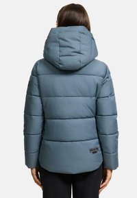 Blå puffjacka med horisontella quiltade sektioner, en stor huva, och en liten logotyp på nedre vänstra sidan. Mjuk, slät textur.