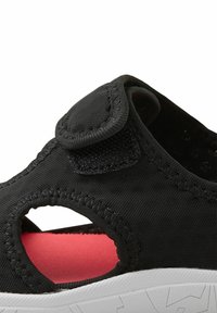 Sandalia negra con parte superior de tela, recortes circulares y tira de velcro. El interior presenta un color rojo en contraste; suela de goma texturizada para mayor agarre.