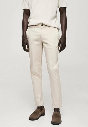 Hombre vestido con pantalones beige ajustados, una camisa de manga corta en beige claro y mocasines de ante marrón, de pie contra un fondo liso.