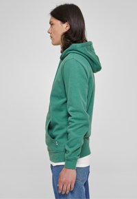 Felpa verde in pile con cappuccio regolabile, tasca a marsupio, e polsini e orlo a costine. Indossata sopra una camicia bianca e abbinata a jeans blu.