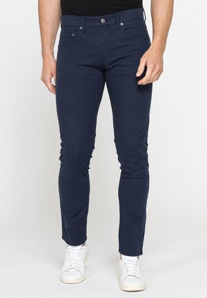 Uomo che indossa pantaloni slim fit blu navy e sneakers bianche, in piedi davanti a uno sfondo chiaro e uniforme.