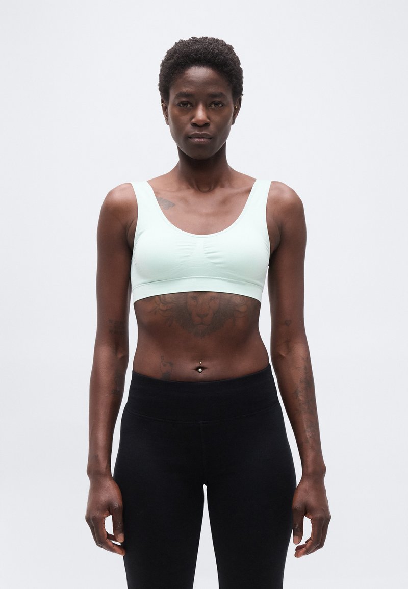 Ευθυγραμμισμένο άτομο που φοράει ανοιχτό πράσινο αθλητικό σουτιέν και μαύρο κολάν, στέκεται με το πρόσωπο προς τα εμπρός με τατουάζ στα χέρια και στον κορμό, μπροστά σε απλό φόντο.