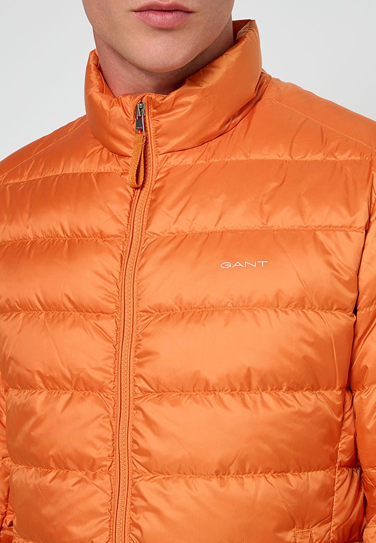 Orangenes Puffermantel mit Reißverschluss und hohem Kragen. Verfügt über gesteppte Abschnitte und ein "GANT"-Logo auf der Brust. Glattes, glänzendes Material.