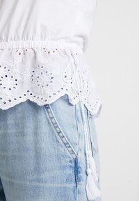 Vit bomullstopp med broderad blommig nederdel, med en scallop-edge och snörning med tofsar. Kombineras med ljusblå denimshorts.