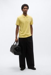 Κίτρινο πόλο πουκάμισο με κοντά μανίκια, κουμπιά στο εμπρόσθιο μέρος και μικρό τυρκουάζ λογότυπο· συνδυασμένο με άνετο μαύρο παντελόνι και υφασμάτινη μαύρη τσάντα.