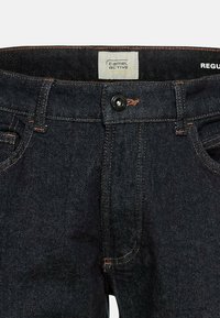 Ciemnoniebieskie dżinsy z zapięciem na guzik, pomarańczowymi szwami i klasycznym pięciokomorowym wzorem. Metka z napisem "Camel Active" w prostokątnej łatce.