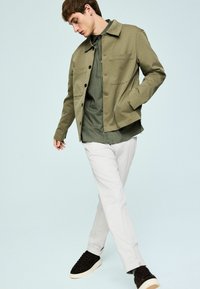 Veste texturée vert olive avec col, deux poches avant, portée sur une chemise à carreaux foncés, associée à un pantalon clair et des baskets noires.