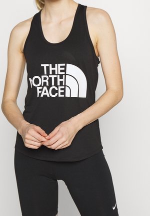 Kobieta w czarnym, bezrękawnikowym topie z dużym białym logo "THE NORTH FACE" i czarnych szortach Nike, z rękami splecionymi przed sobą.