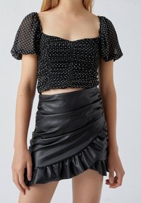 Schwarzes, kurz geschnittenes Oberteil mit Puffärmeln und gepunktetem Muster, mit geraffter Textur. Kombiniert mit einem schwarzen Minirock aus Leder, der gerafft ist und einen Rüschensaum hat.