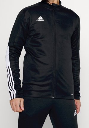 Homme portant une veste zippée noire Adidas avec des bandes blanches sur les manches et un pantalon Adidas noir assorti.