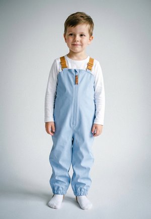 Young child standing wearing light blue overalls with front zipper and brown straps, white long-sleeve shirt, and white socks.