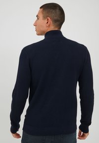 Vīrietis, kurš stāv ar muguru, ģērbies strukturētā tumši zila turtlenecka džemperī un tumšos džinsos uz vienkārša gaiša fona.