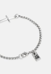 Sidabro grandinėlė su užsegimu, turinti stačiakampį pakabuką su užrašu "IKONA." Lygi tekstūra ir poliruotas paviršius.