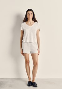 Jauna sieviete ar garām brūnām matiem, valkājot baltu īsroku V-veida kakla topu, gaiši pelēkus šortus un melnas slip-on kurpes, stāvot pie bēša sienas.