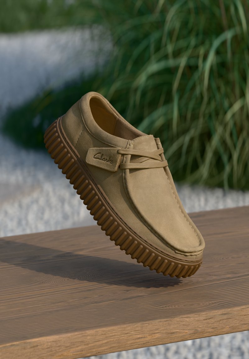 Beige suède schoen met een ronde neus, vetersluiting en geribbelde bruine rubberen zool, geplaatst op een houten oppervlak.