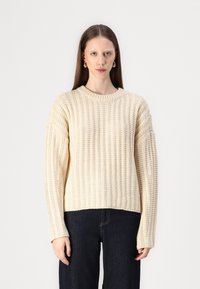 Cream-colored chunky knit sweater with ribbed texture, rounded neckline, and oversized sleeves, paired with dark jeans.
