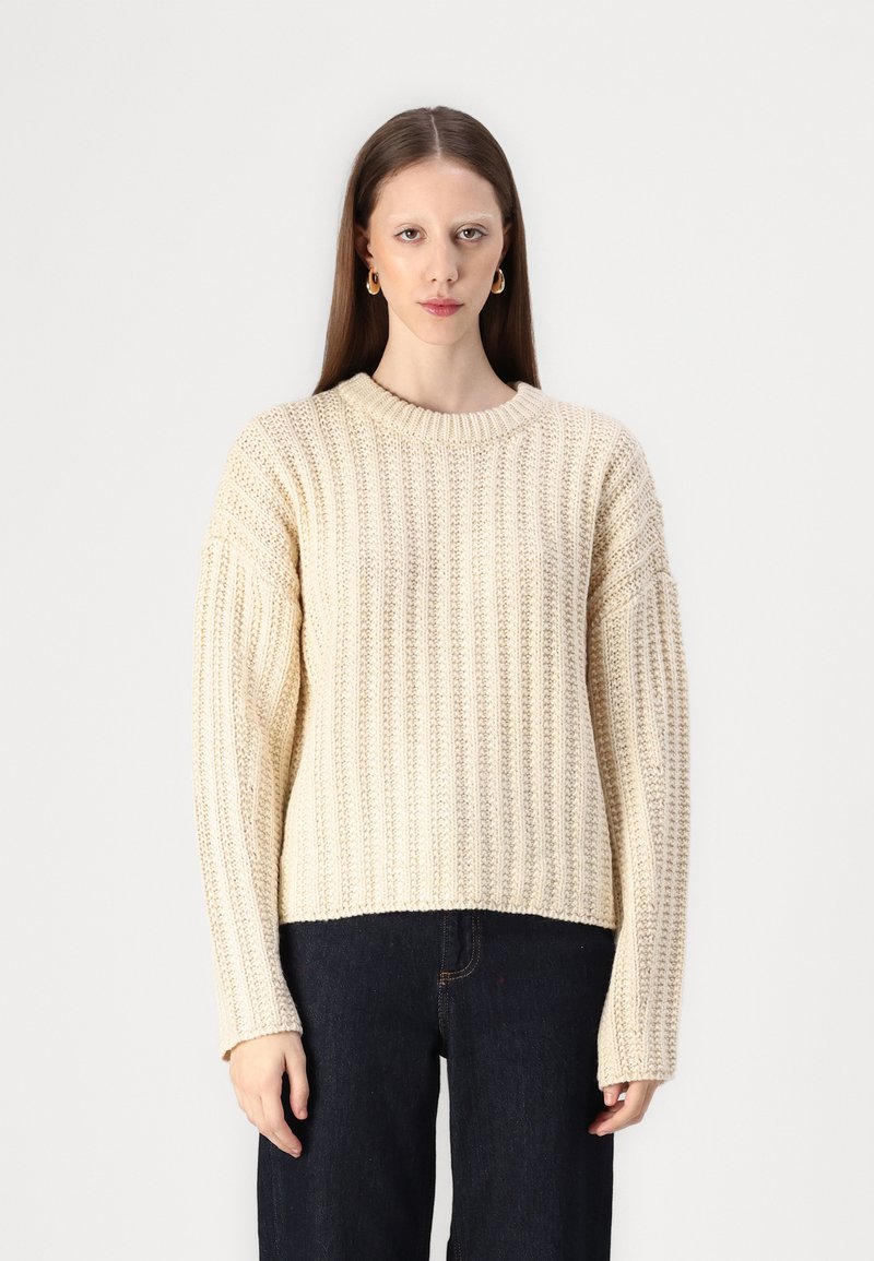 Cream-colored chunky knit sweater with ribbed texture, rounded neckline, and oversized sleeves, paired with dark jeans.