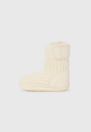 Botte de bébé tricotée couleur crème avec texture côtelée, semelle souple et sangle ajustable autour de la cheville pour un maintien sécurisé.