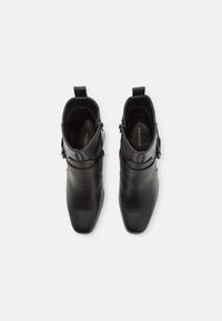 Botas de tobillo de cuero negro con punta afilada, que cuentan con detalles de hebilla y una textura lisa. El diseño es elegante y aerodinámico.