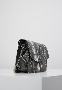 Bolso de mano con estampado de serpiente en blanco y negro, superficie texturizada, cierre de solapa y correa metálica. Forma rectangular, diseño estructurado.