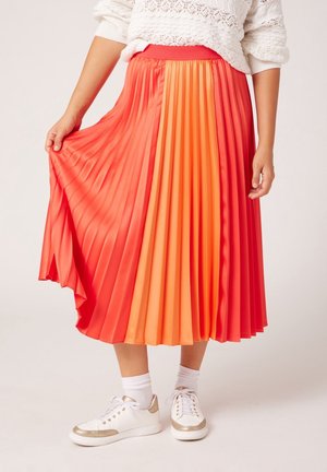 Osoba ubrana w plisowaną spódnicę midi w kolorze czerwono-pomarańczowym, biały sweter, białe skarpetki i białe trampki ze złotymi detalami.