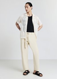 Camicia a maniche corte color panna con motivo a pizzo sopra una canotta nera, abbinata a pantaloni ampi color crema e sandali neri a ciabatta.