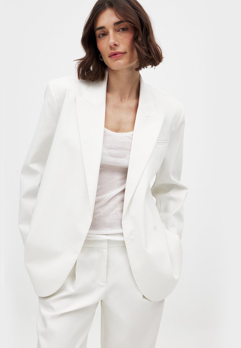Blazer bianco su misura con rever a notched, taschino sul petto singolo e vestibilità rilassata. Indossato sopra una blusa leggera e semitrasparente. Texture liscia.
