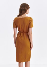 Musztardowa, do kolan, sukienka z krótkimi rękawami, dopasowana w talii i zapinana z tyłu na zamek. Gładki materiał z delikatnym naciągiem i bocznymi kieszeniami.
