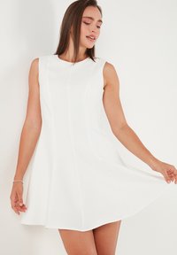 Woman in sleeveless, white A-line dress holding the skirt slightly, standing against a plain light wall.