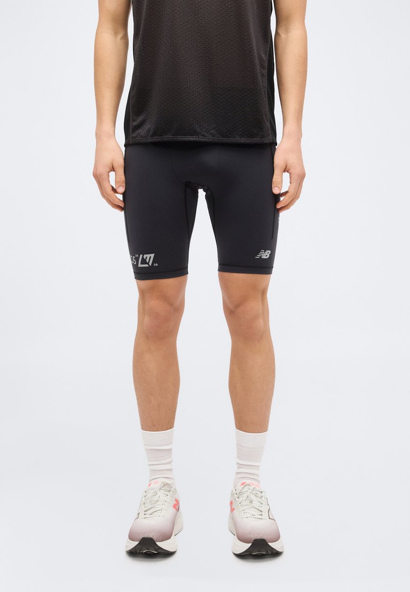Person iført sorte atletikshorts, en sort mesh sportstrøje, hvide sokker og hvide løbesko stående mod en ensfarvet baggrund.