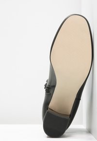 Chaussure en cuir noir avec une finition lisse, dotée d'une semelle plate, d'accents texturés et d'un design sans couture. La partie inférieure est beige.