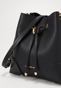 Bolso tipo cubo de cuero negro con textura granulada, detalles en hardware dorado, correa ajustable y cierre de cordón. Presenta un detalle del logo en la parte frontal.