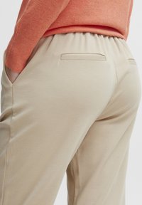 Pantalons beiges avec taille élastique, dotés de poches latérales et de poches arrière discrètes, fabriqués en tissu lisse et léger.
