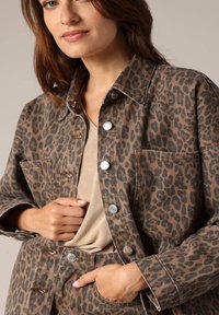 Veste en jean imprimée léopard avec des boutons-pression argentés et deux poches avant, associée à un haut beige. Tissu texturé et design décontracté.