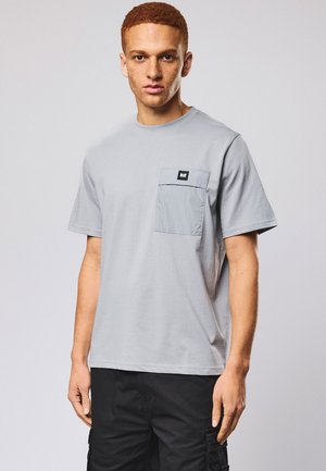 Ung mand med kort rødt hår iført en lysegrå T-shirt med en brystlomme og sorte cargobukser mod en ensfarvet baggrund.