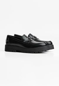 Zwarte leren platformloafers met dikke rubberen zolen en een pennybanddetail, afgebeeld tegen een witte achtergrond.