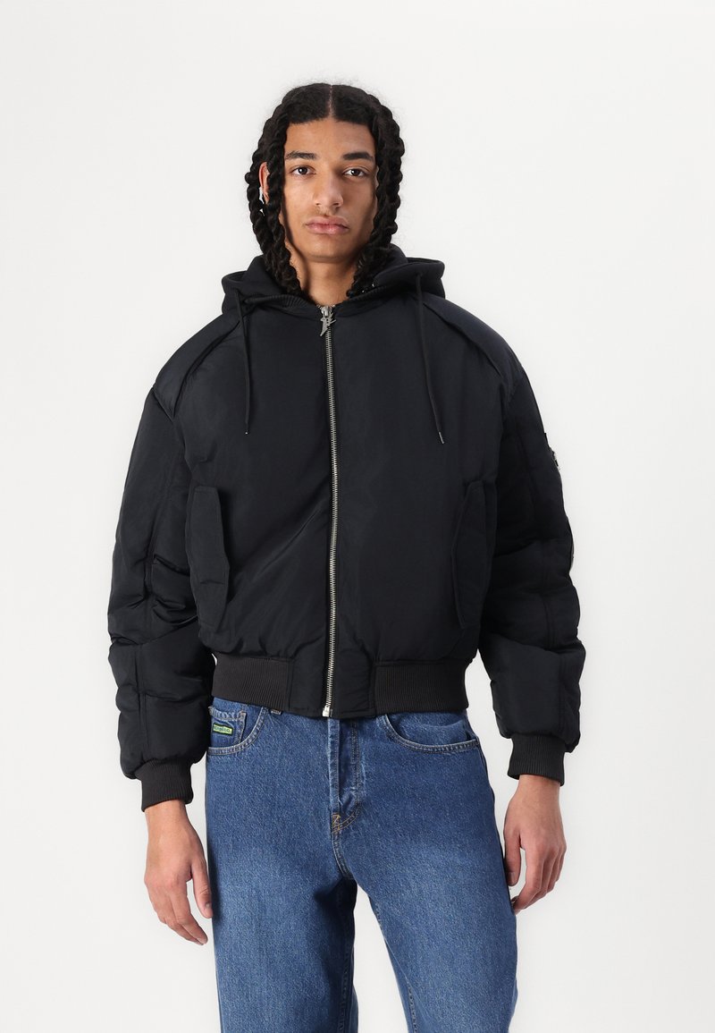 Black puffer jacket with a hood, featuring a zip closure, ribbed cuffs, and side pockets. Smooth fabric with a quilted texture.