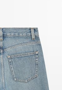 Poche arrière et passants de ceinture d'un jean en denim bleu clair, montrant une couture détaillée et la texture du tissu sur fond blanc uni.