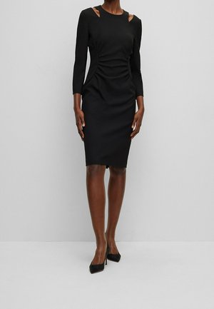 Kobieta w dopasowanej czarnej sukience do kolan z wycięciami na ramionach i rękawami o długości do łokcia, w połączeniu z czarnymi szpilkami.