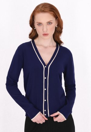 Navy blue cardigan with pearl buttons, V-neckline, and contrasting light beige trim along the edges and cuffs. Soft knit texture.