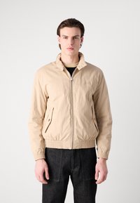 Chaqueta ligera beige con cierre delantero, dos bolsillos laterales, puños elásticos y dobladillo. Lleva puesta sobre una blusa negra y pantalones de mezclilla oscuros.