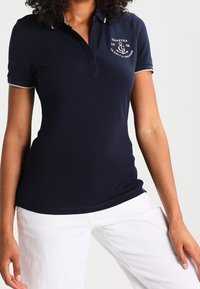 Polo shirt bleu marine en coton, avec une patte de boutonnage à trois boutons, un col et des manches contrastants, et un logo sur la poitrine. Associé à un pantalon blanc.