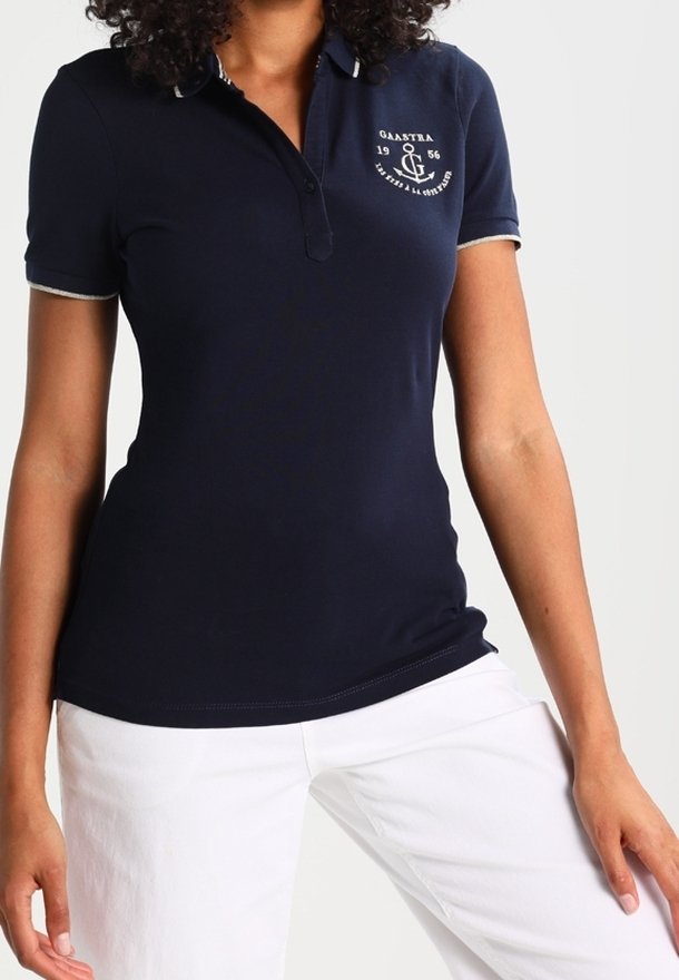 Polo shirt bleu marine en coton, avec une patte de boutonnage à trois boutons, un col et des manches contrastants, et un logo sur la poitrine. Associé à un pantalon blanc.