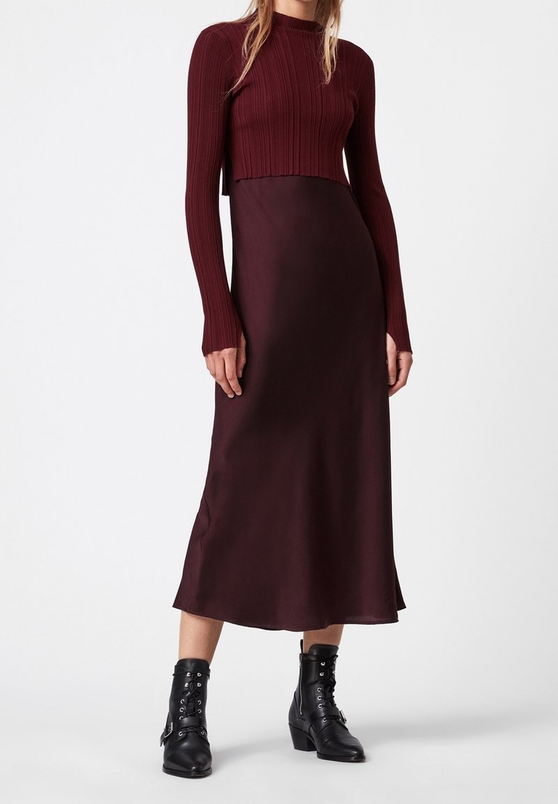Femme portant un long pull côtelé bordeaux et une jupe midi satinée assortie, avec des bottines noires à lacets, sur fond blanc.