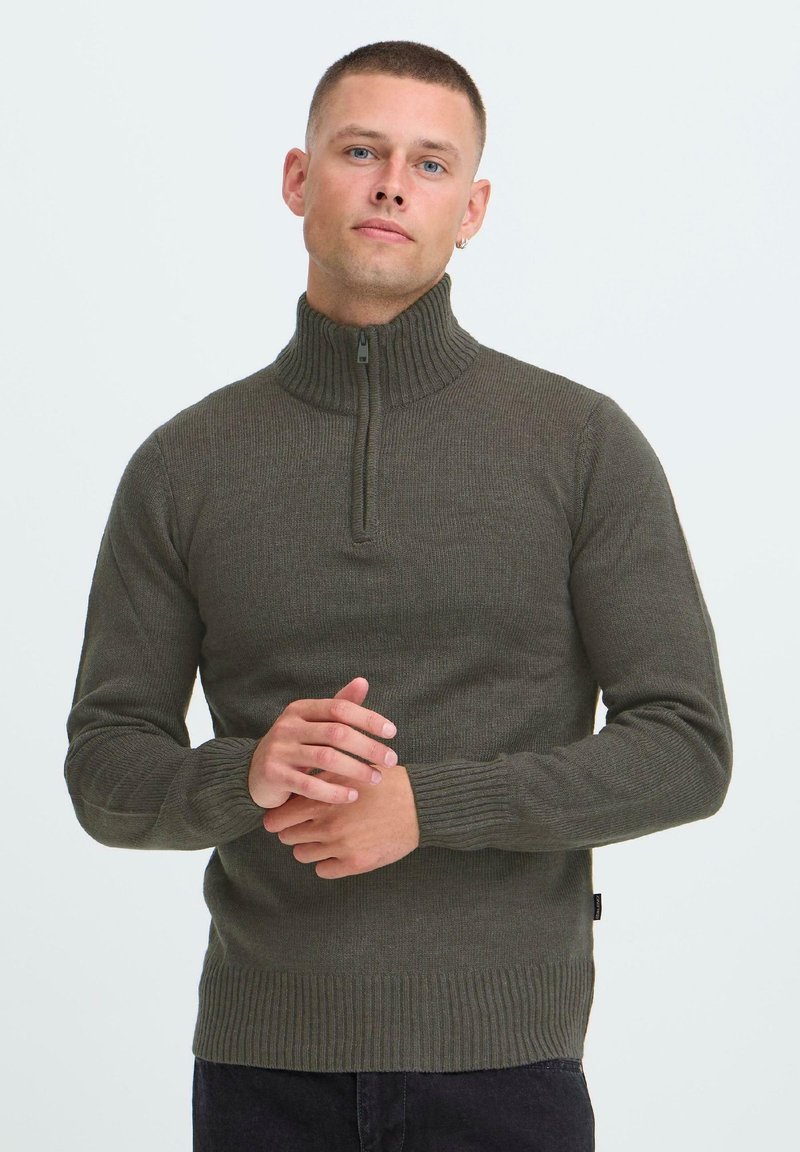 Man met kort haar, gekleed in een donkergroene geribbelde ritssweater, staande tegen een effen lichte achtergrond met de handen samengevouwen.